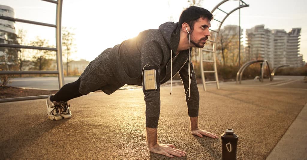 Adult man doing push-ups outdoors with earphones during sunrise, showcasing fitness and healthy lifestyle.