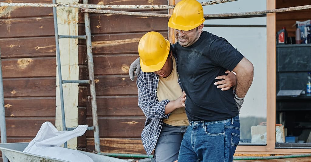 Construction workers wearing helmets assist an injured colleague at a building site with wooden cabins.