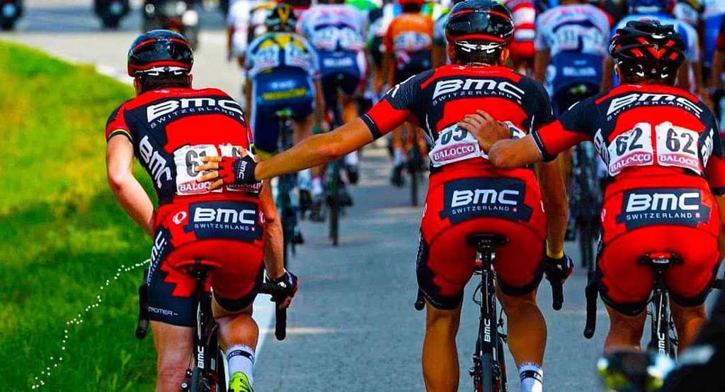 "Cyclists racing on an indoor velodrome track, wearing colorful jerseys and helmets. The riders are using fixed-gear track bikes with drop handlebars, leaning forward in an aerodynamic position. The wooden track features blue and red guideline markings. Banners and advertisements are visible in the background, indicating an organized cycling event.