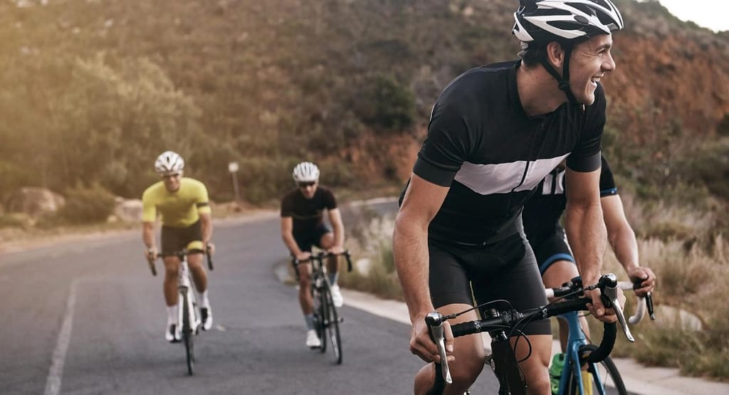 Wayne State Cycle Track. "Cyclists racing on an indoor velodrome track, wearing colorful jerseys and helmets. The riders are using fixed-gear track bikes with drop handlebars, leaning forward in an aerodynamic position. The wooden track features blue and red guideline markings. Banners and advertisements are visible in the background, indicating an organized cycling event.