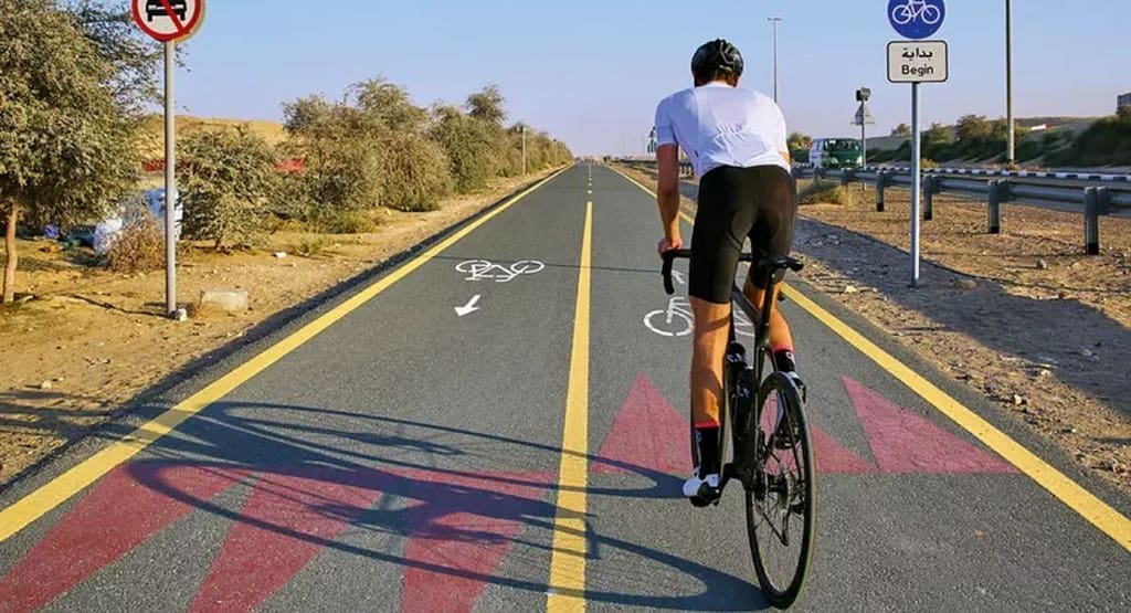 "Cyclists racing on an indoor velodrome track, wearing colorful jerseys and helmets. The riders are using fixed-gear track bikes with drop handlebars, leaning forward in an aerodynamic position. The wooden track features blue and red guideline markings. Banners and advertisements are visible in the background, indicating an organized cycling event.