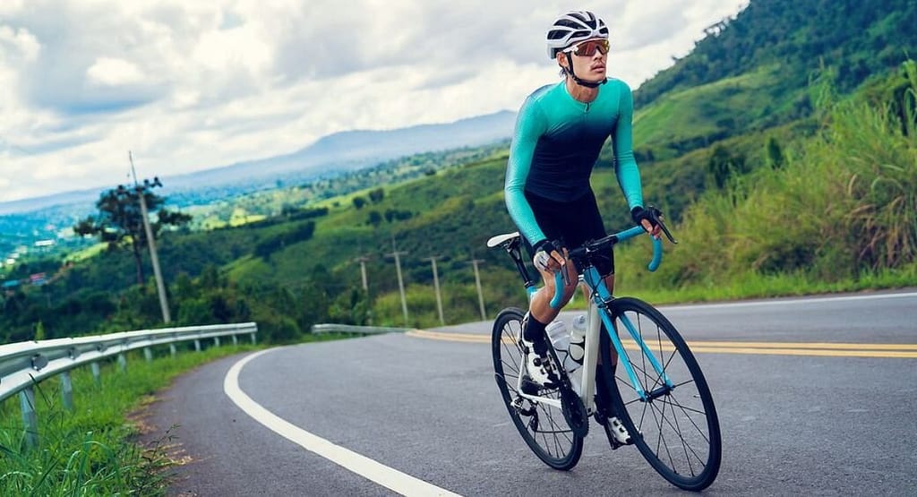 Impact Cycle Data Analytics. A cyclist wearing a teal and black aerodynamic outfit rides a road bike uphill on a winding mountain road. The background features lush green hills, scattered trees, and a partly cloudy sky. The cyclist is equipped with a helmet, sunglasses, and gloves, appearing focused as they pedal along the smooth asphalt road with a guardrail on one side.