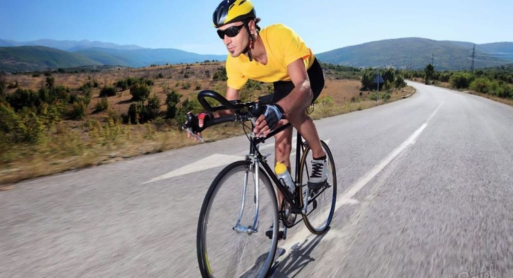 A cyclist wearing a teal and black aerodynamic outfit rides a road bike uphill on a winding mountain road. The background features lush green hills, scattered trees, and a partly cloudy sky. The cyclist is equipped with a helmet, sunglasses, and gloves, appearing focused as they pedal along the smooth asphalt road with a guardrail on one side.