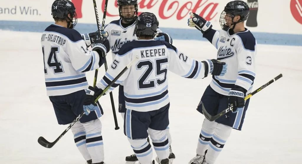 A University of Maine ice hockey goaltender in a blue and black uniform with the number 1, standing in front of the net in a ready position. The player is wearing full protective gear, including a helmet with a cage, large leg pads, a blocker, and a catching glove. The background features the hockey rink and goalposts. UMaine Hockey
