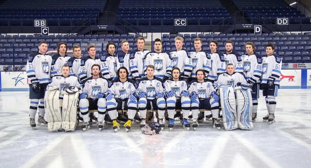UMaine Hockey A University of Maine ice hockey goaltender in a blue and black uniform with the number 1, standing in front of the net in a ready position. The player is wearing full protective gear, including a helmet with a cage, large leg pads, a blocker, and a catching glove. The background features the hockey rink and goalposts.