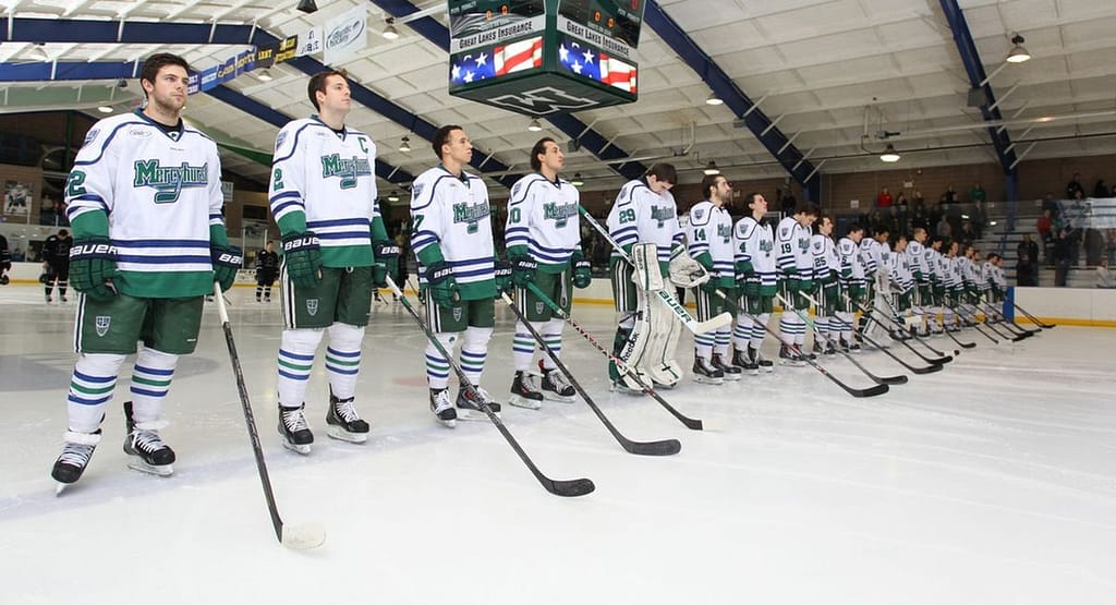 UMaine Hockey A University of Maine ice hockey goaltender in a blue and black uniform with the number 1, standing in front of the net in a ready position. The player is wearing full protective gear, including a helmet with a cage, large leg pads, a blocker, and a catching glove. The background features the hockey rink and goalposts.