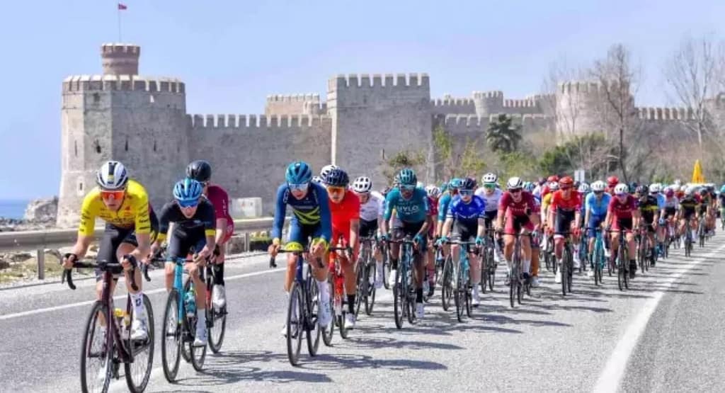"United Cycling race competitors sprint toward the finish line, with the leading cyclist in a yellow 'LEADER' jersey raising his arm in victory. The pack of riders follows closely behind on a winding road with patches of snow in the background, showcasing the intensity of the competition."