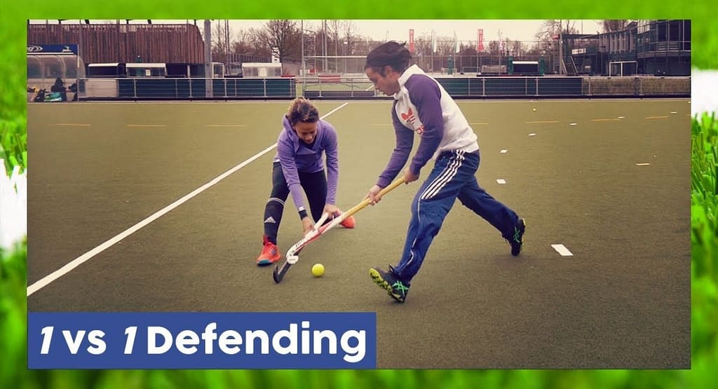 Two female players in an Arts Academy Field Hockey match compete for possession of the ball on a bright green artificial turf field. One player, wearing a black and yellow uniform, skillfully controls the ball with her hockey stick, while the other, in a light blue uniform, closely defends. Other players and banners are visible in the background, adding to the competitive atmosphere of the game.