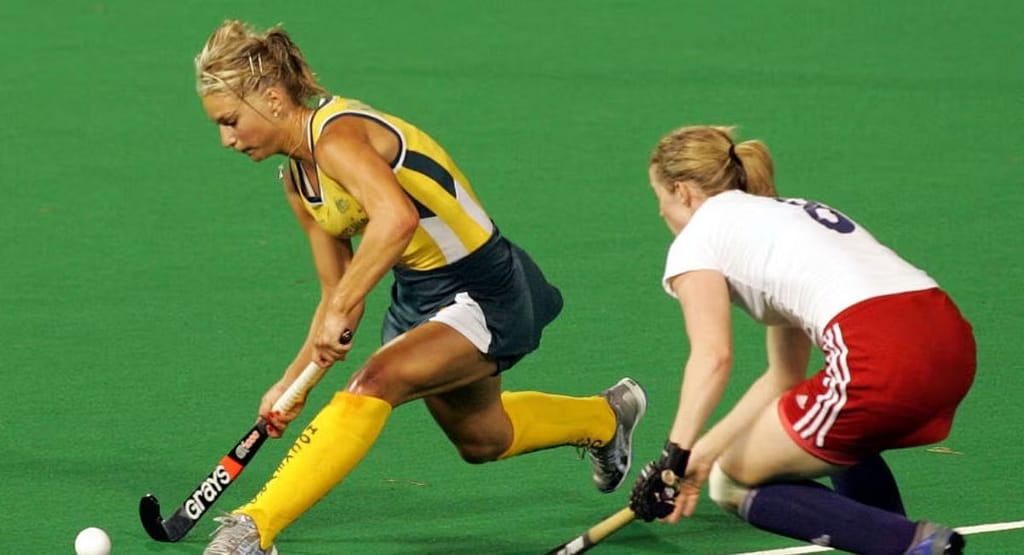 Two female players in an Arts Academy Field Hockey match compete for possession of the ball on a bright green artificial turf field. One player, wearing a black and yellow uniform, skillfully controls the ball with her hockey stick, while the other, in a light blue uniform, closely defends. Other players and banners are visible in the background, adding to the competitive atmosphere of the game.