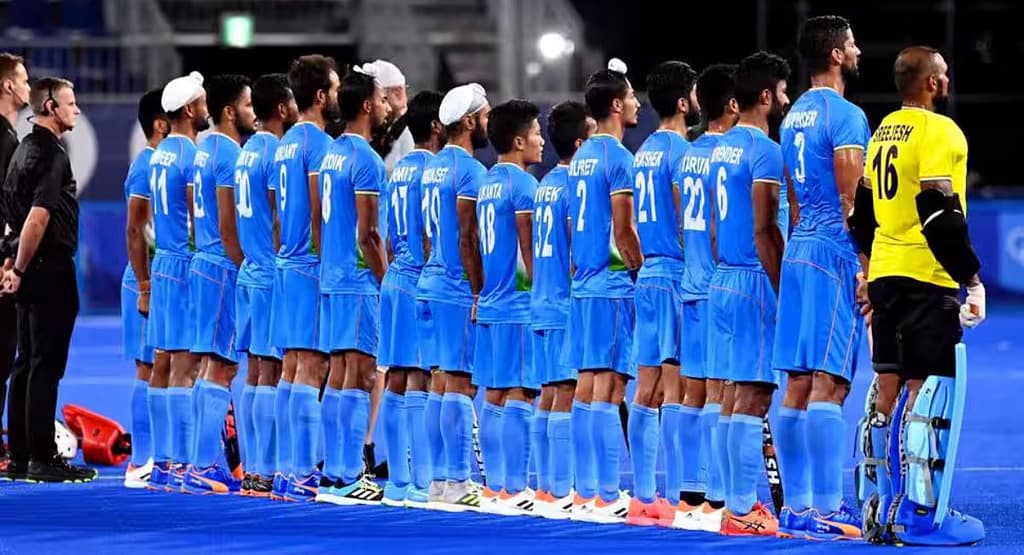 Two female players in an Arts Academy Field Hockey match compete for possession of the ball on a bright green artificial turf field. One player, wearing a black and yellow uniform, skillfully controls the ball with her hockey stick, while the other, in a light blue uniform, closely defends. Other players and banners are visible in the background, adding to the competitive atmosphere of the game.