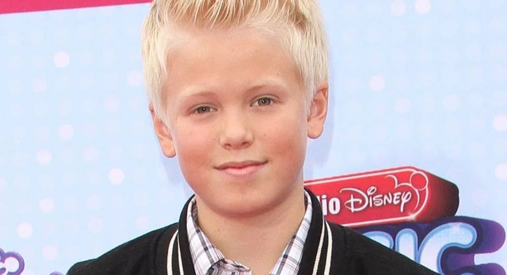 "Bremen High School Football Injury: A soccer player in a blue jersey lies on the artificial turf field, appearing to have sustained a leg injury. A coach or medical staff member kneels beside them, holding their leg to assess or provide first aid. A soccer ball is nearby, and in the background, players in orange jerseys continue the game."