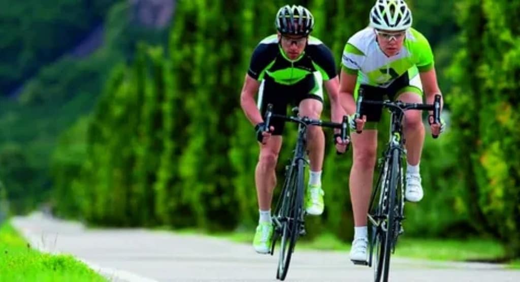 "A cyclist wearing black cycling gear is riding a high-performance road bike indoors, mounted on a smart trainer. The setting features a dark, modern training space with ambient lighting, a circular LED light on the wall, and cycling-related decor, creating an immersive workout environment." Cycle Training