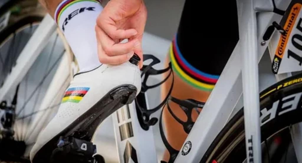 A close-up of a cyclist's foot wearing high-performance white cycling shoes with dial fasteners, clipped into the pedal of a road bike. The background features a blurred bicycle wheel and a sunlit outdoor setting, emphasizing the sport of cycling and professional gear."
Let me know if you'd like any modifications!