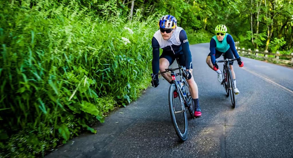 "A cyclist wearing black cycling gear is riding a high-performance road bike indoors, mounted on a smart trainer. The setting features a dark, modern training space with ambient lighting, a circular LED light on the wall, and cycling-related decor, creating an immersive workout environment." Cycling: When to Take a Rest Week