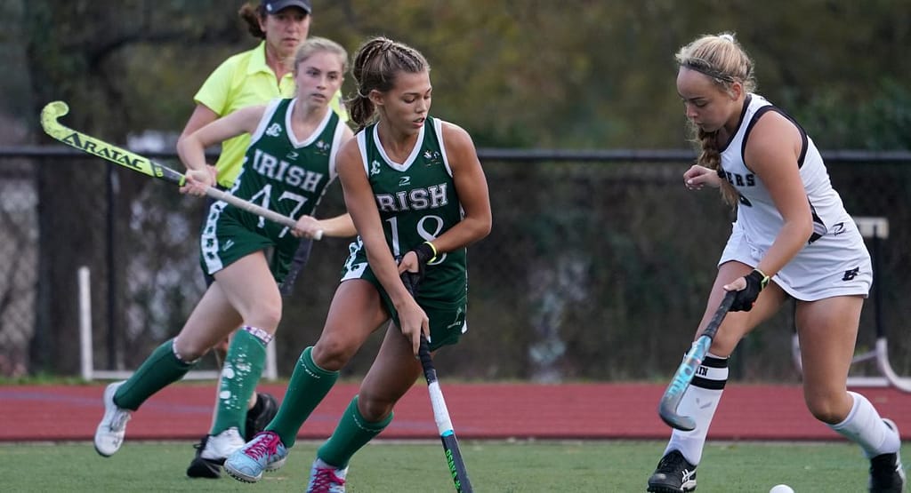 A competitive moment in a Jazmin Field Hockey match, featuring a player in a green and white uniform skillfully maneuvering the ball past a goalkeeper in full protective gear. The intensity of the game is evident as both athletes focus on the play, highlighting the excitement and determination in field hockey.