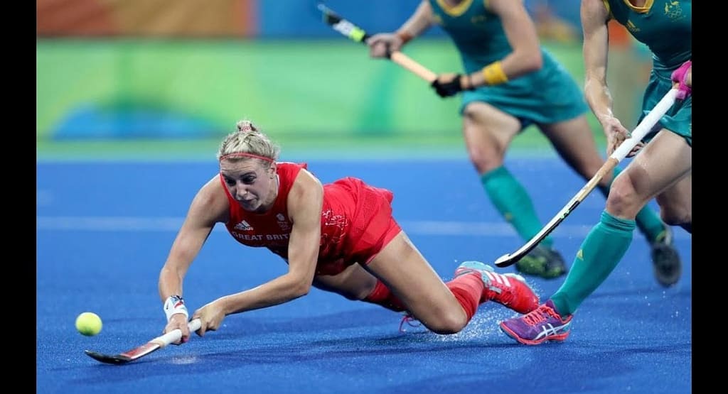 A competitive moment in a Jazmin Field Hockey match, featuring a player in a green and white uniform skillfully maneuvering the ball past a goalkeeper in full protective gear. The intensity of the game is evident as both athletes focus on the play, highlighting the excitement and determination in field hockey.