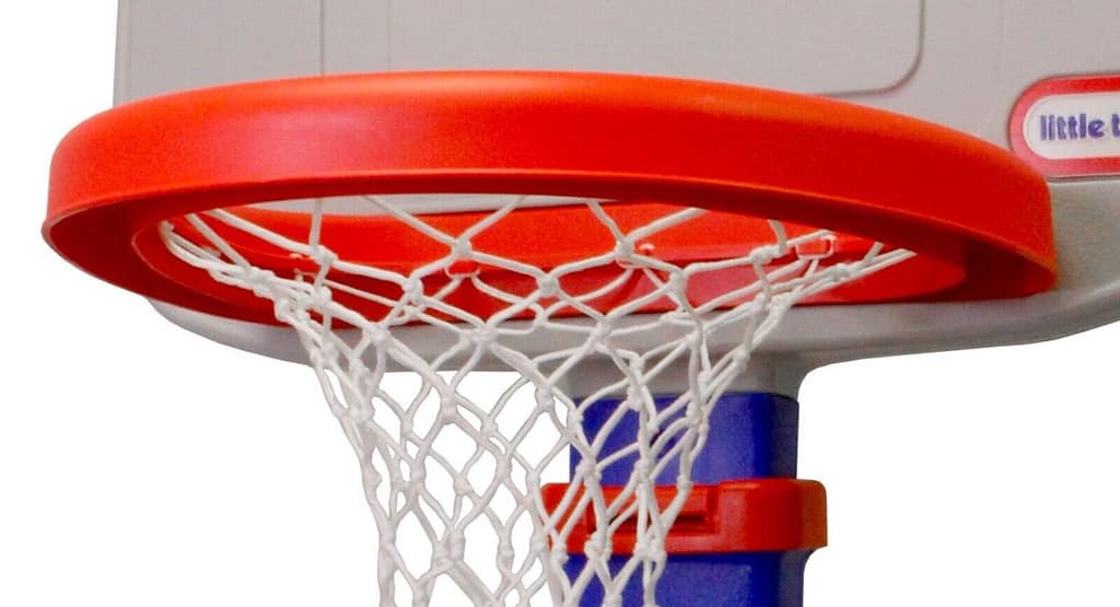 Two young boys playing with a Little Tikes Basketball Hoop outdoors on a sunny day. One child is smiling with his hands raised, while the other is attempting a shot with an orange basketball. The colorful plastic hoop, with a blue stand and red rim, is set against a backdrop of green hills and a clear blue sky, highlighting a fun, active playtime scene.