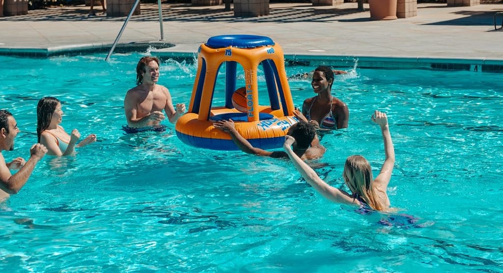 A group of four people enjoying a game with a Pool Basketball Hoop in a backyard swimming pool. Two men are jumping to shoot and block the basketball, while a woman in a pink shirt and another player in a black shirt look on excitedly. The adjustable Pool Basketball Hoop is positioned at the pool's edge, with a water slide and lush greenery in the background, capturing a fun and energetic summer activity.