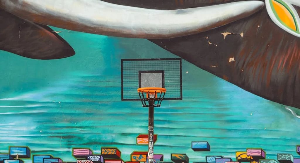 A group of four people enjoying a game with a Pool Basketball Hoop in a backyard swimming pool. Two men are jumping to shoot and block the basketball, while a woman in a pink shirt and another player in a black shirt look on excitedly. The adjustable Pool Basketball Hoop is positioned at the pool's edge, with a water slide and lush greenery in the background, capturing a fun and energetic summer activity.