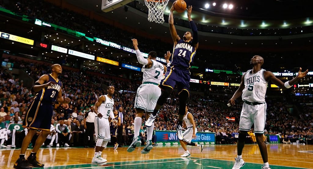 "Basketball player in a white jersey dribbling past defenders during a competitive indoor game, with spectators watching from the stands representing the intensity and skill seen in MetroWest Basketball competitions."