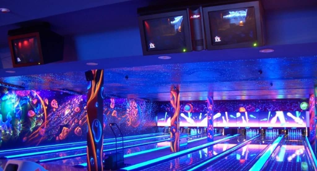 “Wide shot of a glow-in-the-dark bowling lane under ultraviolet lights, with neon lane markings and pins glowing bright pink at the far end.”