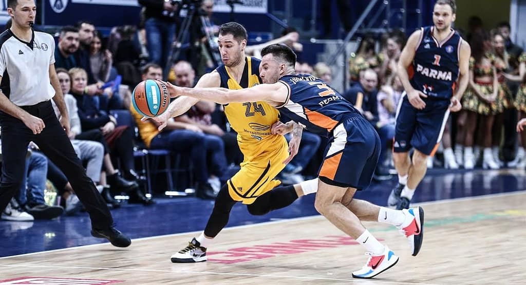 "Basketball player in a white jersey dribbling past defenders during a competitive indoor game, with spectators watching from the stands representing the intensity and skill seen in MetroWest Basketball competitions."