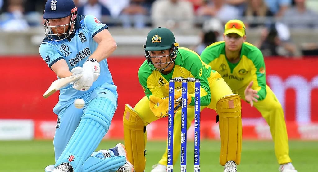 Two cricketers in action during a match one batsman wearing protective gear and mid-swing with a wooden bat, while the wicketkeeper in white and maroon gear is ready behind the stumps. The red cricket ball is captured mid-air against a green field background, highlighting the intensity and skill involved. Represents the emphasis of TCA Cricket on quality gear and professional coaching in modern cricket training.
