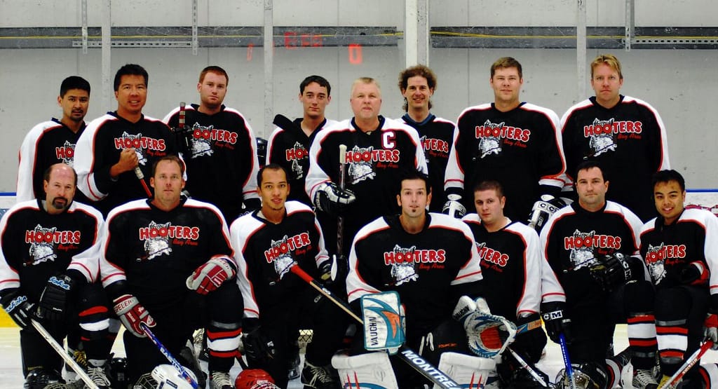 Action shot from a professional ice hockey game showing players in motion. While the image features teams in black and white/red jerseys, it does not depict the classic Chiefs hockey jersey made famous by the fictional Charlestown Chiefs from Slap Shot. The Chiefs jersey typically features blue and gold striping with bold "Chiefs" lettering across the chest