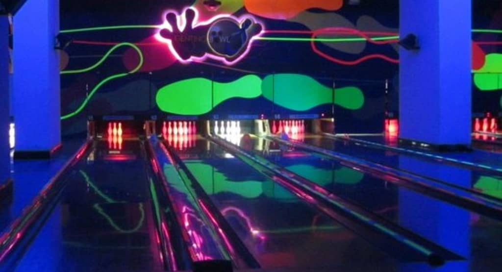 “Wide shot of a glow-in-the-dark bowling lane under ultraviolet lights, with neon lane markings and pins glowing bright pink at the far end.”