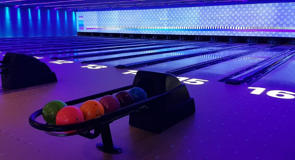 “Wide shot of a glow-in-the-dark bowling lane under ultraviolet lights, with neon lane markings and pins glowing bright pink at the far end.”