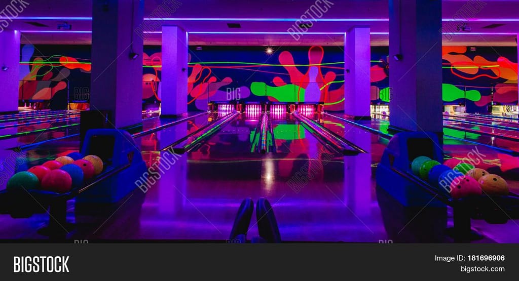 “Wide shot of a glow-in-the-dark bowling lane under ultraviolet lights, with neon lane markings and pins glowing bright pink at the far end.”
