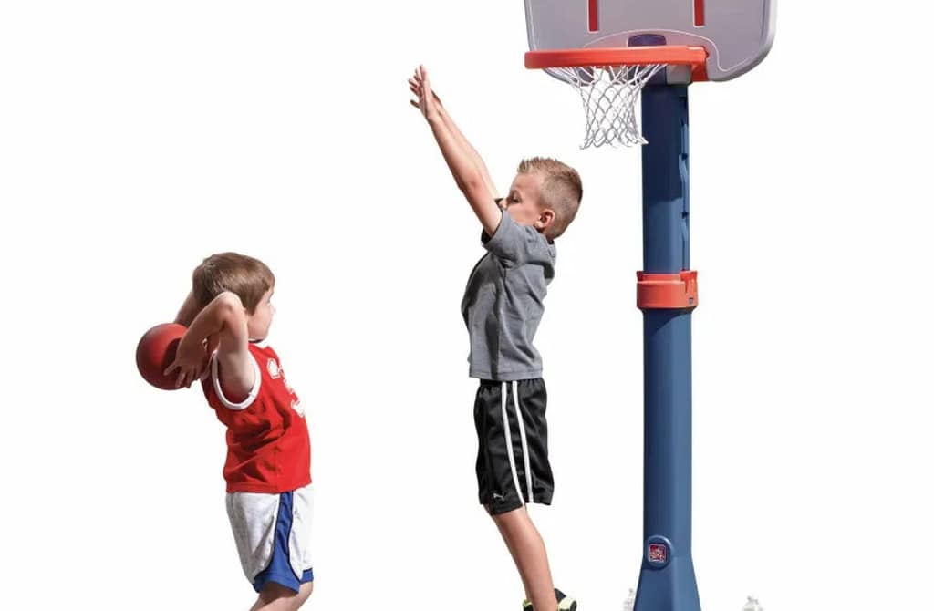 Two young boys playing with a Little Tikes Basketball Hoop outdoors on a sunny day. One child is smiling with his hands raised, while the other is attempting a shot with an orange basketball. The colorful plastic hoop, with a blue stand and red rim, is set against a backdrop of green hills and a clear blue sky, highlighting a fun, active playtime scene.