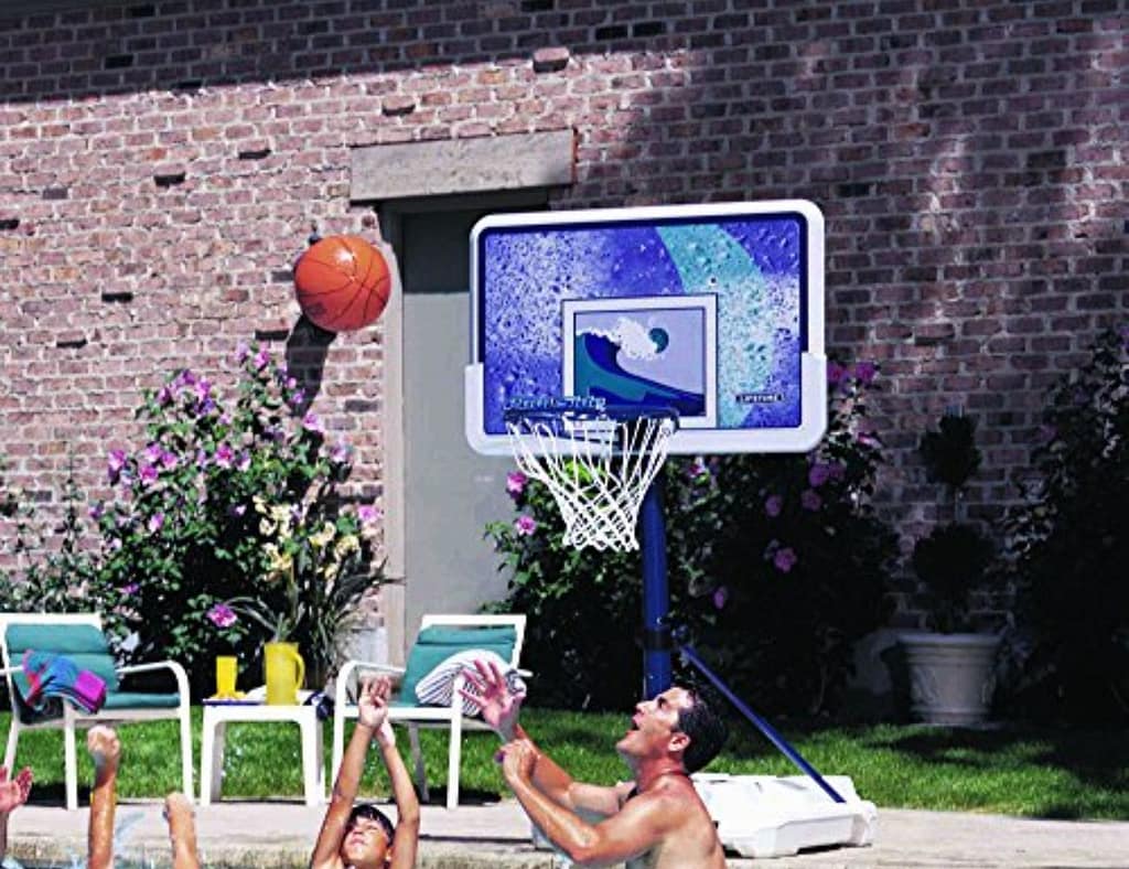 A group of four people enjoying a game with a Pool Basketball Hoop in a backyard swimming pool. Two men are jumping to shoot and block the basketball, while a woman in a pink shirt and another player in a black shirt look on excitedly. The adjustable Pool Basketball Hoop is positioned at the pool's edge, with a water slide and lush greenery in the background, capturing a fun and energetic summer activity.