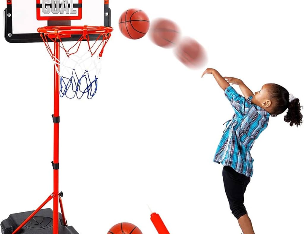Two young boys playing with a Little Tikes Basketball Hoop outdoors on a sunny day. One child is smiling with his hands raised, while the other is attempting a shot with an orange basketball. The colorful plastic hoop, with a blue stand and red rim, is set against a backdrop of green hills and a clear blue sky, highlighting a fun, active playtime scene.
