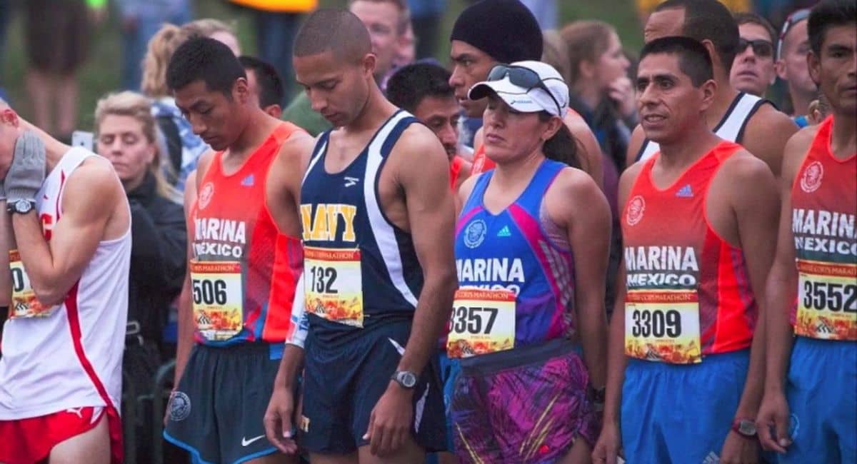 A group of average marathon time for first-time runners standing together at the starting line. The participants are wearing athletic gear, with some sporting shirts labeled "Marina Mexico" and "Navy." Their bib numbers are visible, and they appear focused and prepared for the race. The background features a crowd of spectators.