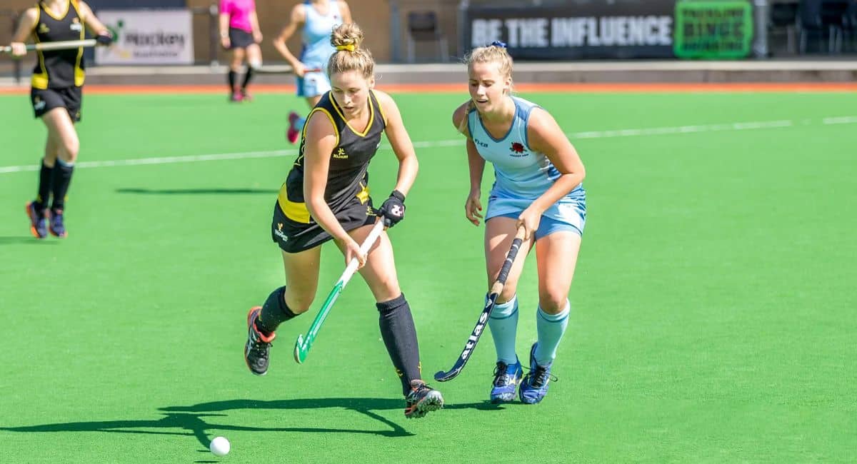 Two female players in an Arts Academy Field Hockey match compete for possession of the ball on a bright green artificial turf field. One player, wearing a black and yellow uniform, skillfully controls the ball with her hockey stick, while the other, in a light blue uniform, closely defends. Other players and banners are visible in the background, adding to the competitive atmosphere of the game.