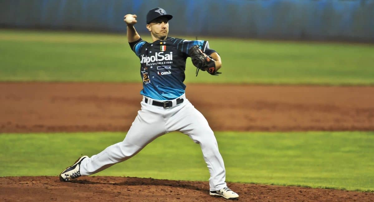 A baseball player mid-pitch on the mound, wearing a black baseball cap paired with a blue and black jersey, white pants, and cleats. The cap complements the player's athletic look while providing functional sun protection, highlighting its dual role as a style and performance essential for 2025.