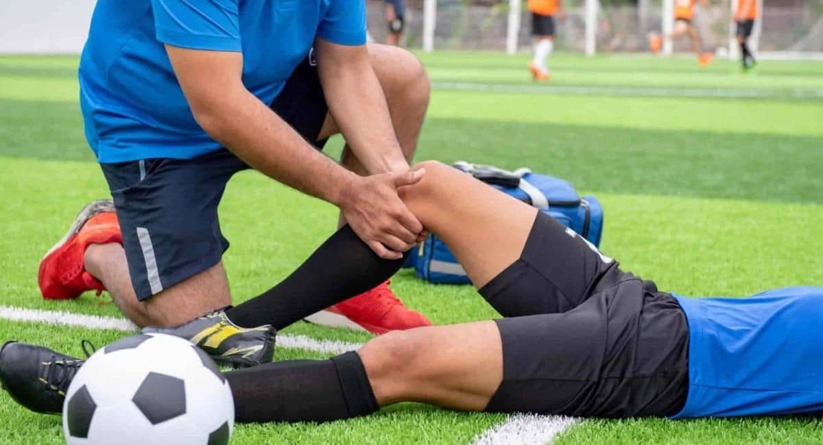 "Bremen High School Football Injury: A soccer player in a blue jersey lies on the artificial turf field, appearing to have sustained a leg injury. A coach or medical staff member kneels beside them, holding their leg to assess or provide first aid. A soccer ball is nearby, and in the background, players in orange jerseys continue the game."