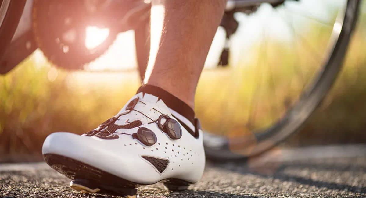 A close-up of a cyclist's foot wearing high-performance white cycling shoes with dial fasteners, clipped into the pedal of a road bike. The background features a blurred bicycle wheel and a sunlit outdoor setting, emphasizing the sport of cycling and professional gear." Let me know if you'd like any modifications!