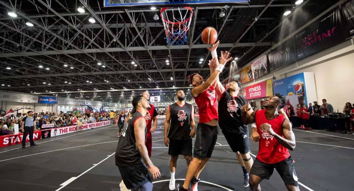 Players compete intensely on an indoor basketball court featuring advanced lighting and modern infrastructure. The venue is packed with spectators, with banners displaying sponsorships from Pepsi and a "Fitness Studio" visible in the background. The court’s sleek design and overhead lighting highlight the role of advanced technologies in enhancing gameplay and the spectator experience.