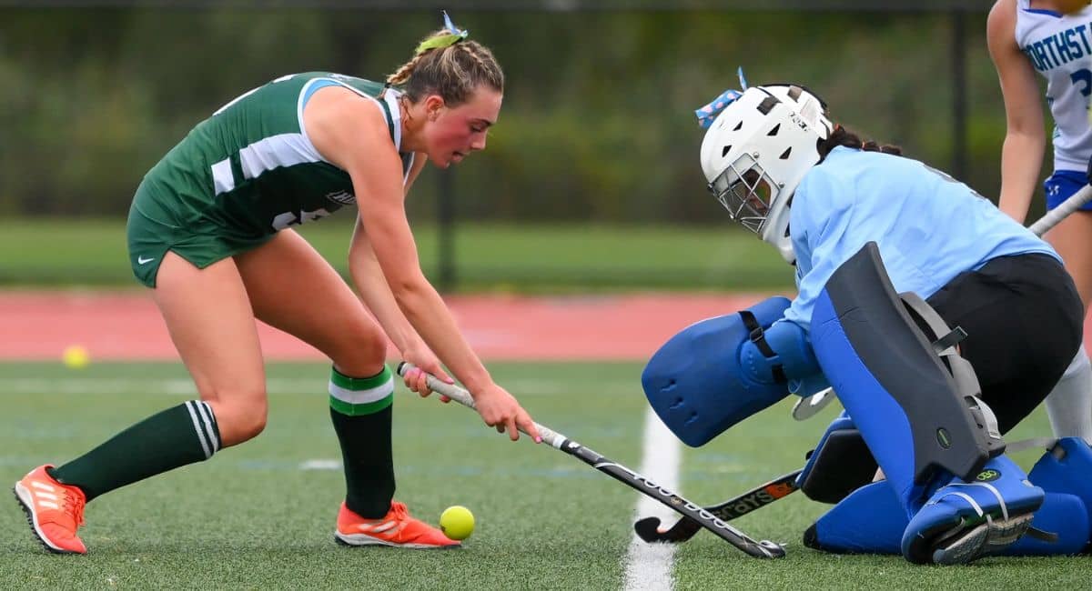 A competitive moment in a Jazmin Field Hockey match, featuring a player in a green and white uniform skillfully maneuvering the ball past a goalkeeper in full protective gear. The intensity of the game is evident as both athletes focus on the play, highlighting the excitement and determination in field hockey.