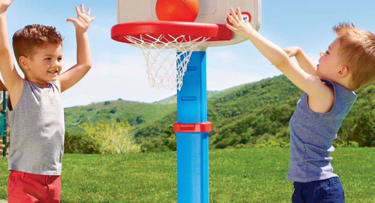 Two young boys playing with a Little Tikes Basketball Hoop outdoors on a sunny day. One child is smiling with his hands raised, while the other is attempting a shot with an orange basketball. The colorful plastic hoop, with a blue stand and red rim, is set against a backdrop of green hills and a clear blue sky, highlighting a fun, active playtime scene.