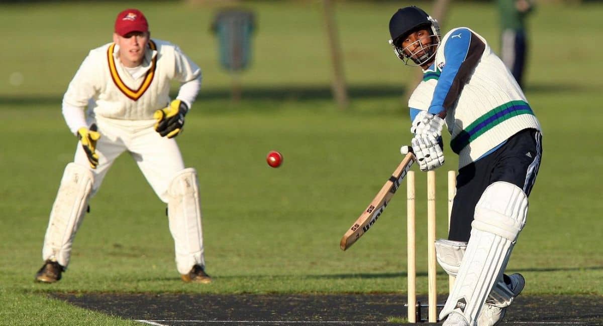 Two cricketers in action during a match one batsman wearing protective gear and mid-swing with a wooden bat, while the wicketkeeper in white and maroon gear is ready behind the stumps. The red cricket ball is captured mid-air against a green field background, highlighting the intensity and skill involved. Represents the emphasis of TCA Cricket on quality gear and professional coaching in modern cricket training.