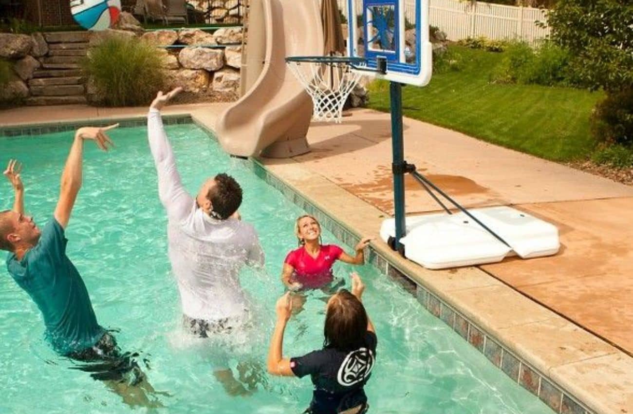 A group of four people enjoying a game with a Pool Basketball Hoop in a backyard swimming pool. Two men are jumping to shoot and block the basketball, while a woman in a pink shirt and another player in a black shirt look on excitedly. The adjustable Pool Basketball Hoop is positioned at the pool's edge, with a water slide and lush greenery in the background, capturing a fun and energetic summer activity.